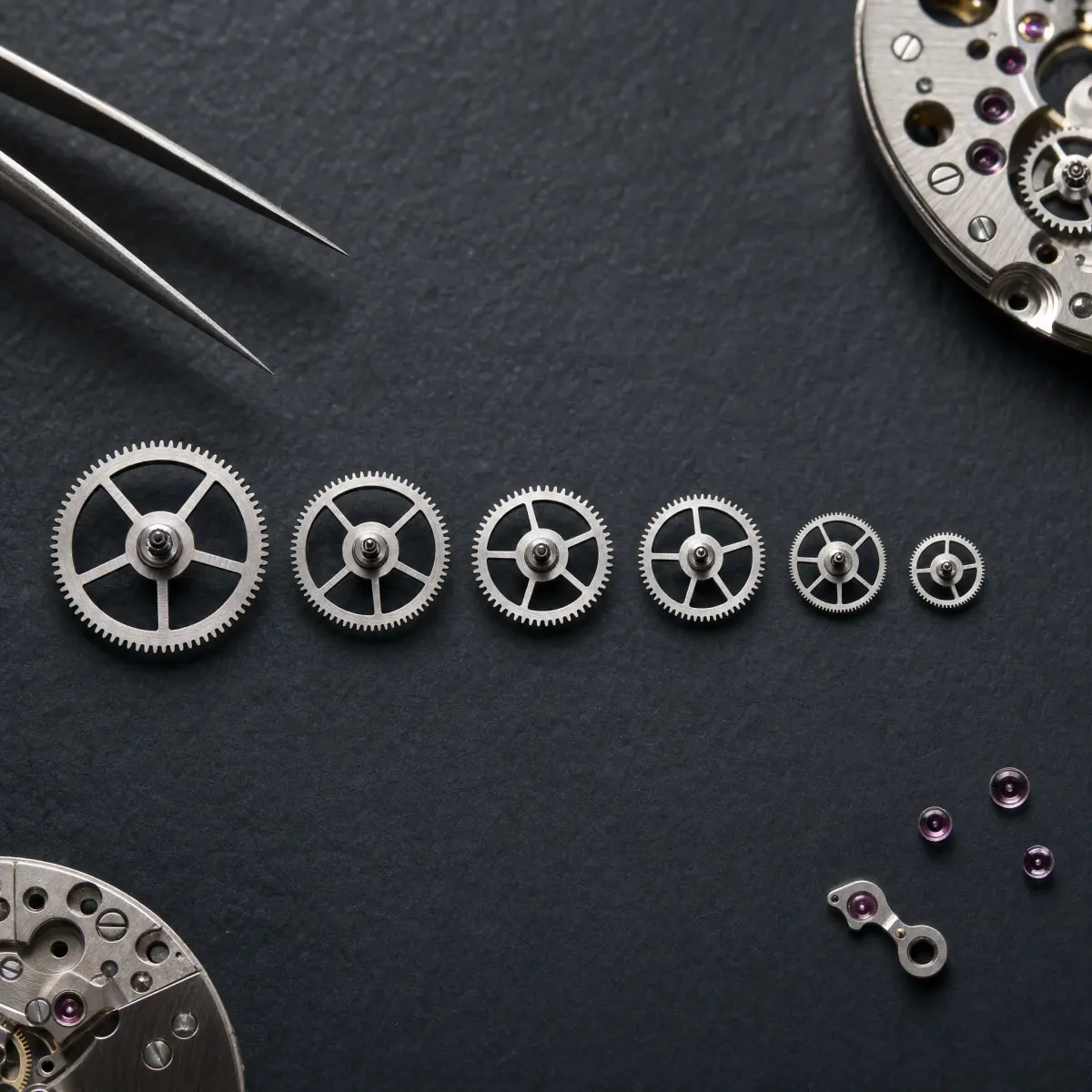 Six polished steel watch gears of different sizes laid out in a row on dark slate, tweezers at the edge.